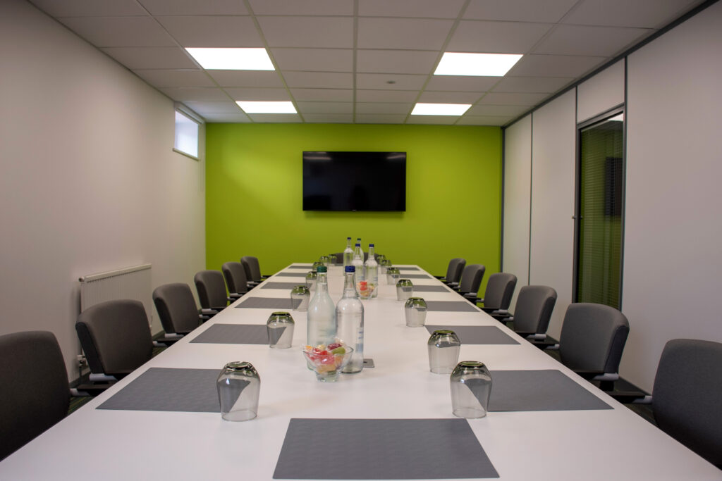Long boardroom style meeting rom for 14 people with a large tv on a green wall.